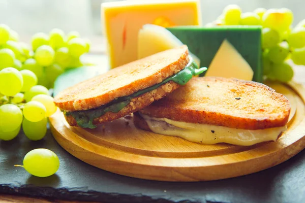 木のテーブルの上に黒スレートのチーズボード上のチーズと緑のブドウと2つのホットグリルや揚げサンドイッチ — ストック写真