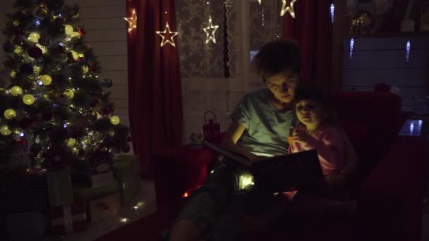 Mamá y su hija leyeron un libro sobre el fondo del árbol de Navidad — Vídeos de Stock