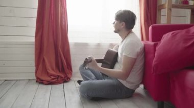 Yerde oturan bir adam evde gitar çalar ve şarkı söyler..