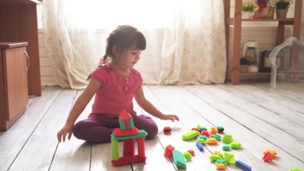 Pequena menina bonita senta-se no chão na sala e brinca com um construtor colorido — Vídeo de Stock
