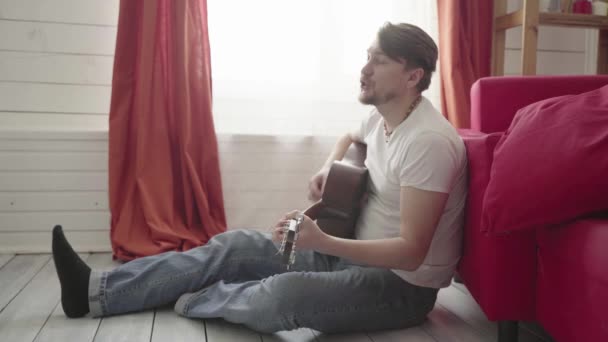 床に座った男が家でギターを弾きながら歌います. — ストック動画