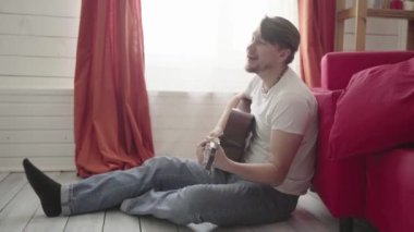 Yerde oturan bir adam evde gitar çalar ve şarkı söyler..