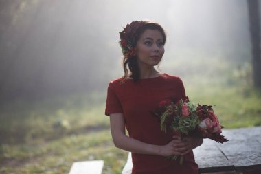 Elinde çiçeklerle bir orman perisi suretinde bir kız. Güneş ışınları ormanda yürüyen kadını dövüyor.