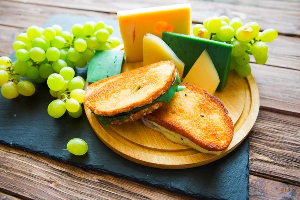 木のテーブルの上に黒スレートのチーズボード上のチーズと緑のブドウと2つのホットグリルや揚げサンドイッチ — ストック写真