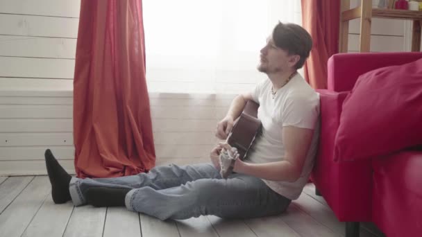 Мужчина сидит на полу, играет на гитаре дома и поет: — стоковое видео