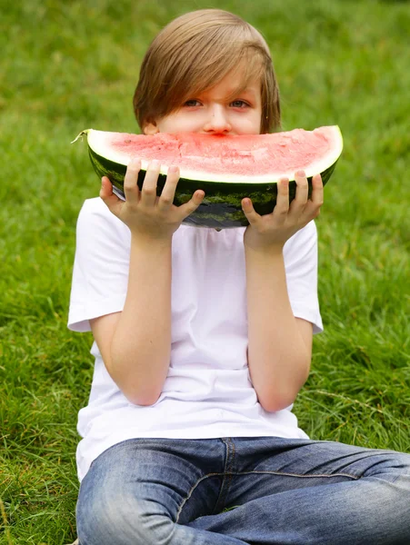 Ładny blond chłopiec zjada dojrzały arbuz czerwonego na zielony łąka — Zdjęcie stockowe