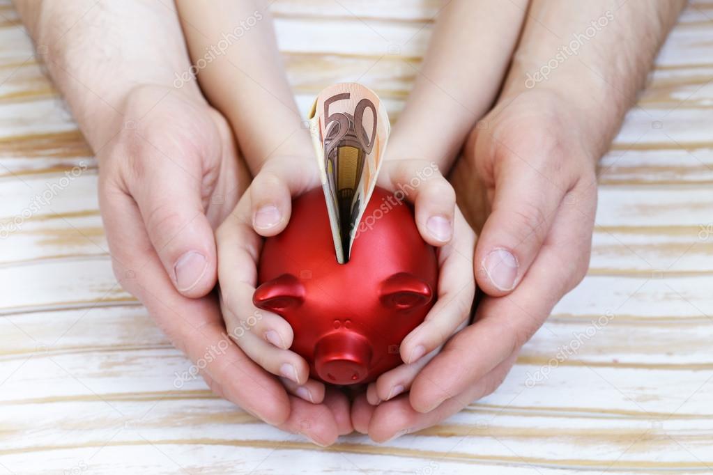 close up of father and son hands holding red piggy bank