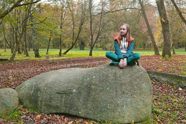 School age girl in turquoise and orange clothes sits on a huge shapeless boulder in an old autumn park, color editing photo