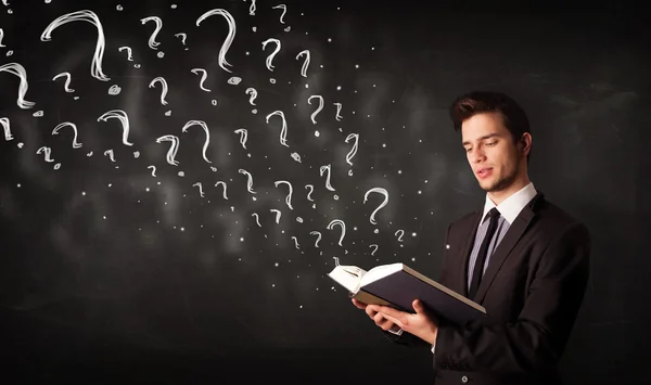 Young man reading a book with question marks coming out from it — Stock Photo, Image