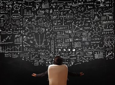 İşadamı elinde çizelgelerle bir tahtanın önünde oturuyor.