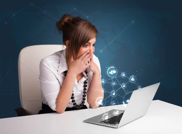Jovencita sentada en el escritorio y escribiendo en el portátil con red social —  Fotos de Stock