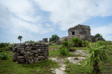 Tulum, Meksika 'da parçalı bulutlu bir gökyüzünün altında verimli tropikal bitki örtüsü ve palmiye ağaçları ile çevrili eski bir Maya tapınağının yıpranmış taş kalıntıları.