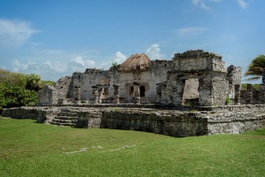 Yıpranmış kireçtaşından duvarlar, sazdan çatıları olan birden fazla yapı ve tülünde mavi gökyüzü altında manikürlü yeşil çimlerle çevrili taş temeller içeren geniş bir Maya arkeolojik alanı..