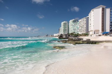 Kristal berrak turkuaz okyanus dalgaları el değmemiş beyaz kuma nazikçe yaklaşırken, çağdaş beyaz otel kuleleri parlak mavi gökyüzünün altında manzaralı kıyı şeridi boyunca uzanır..