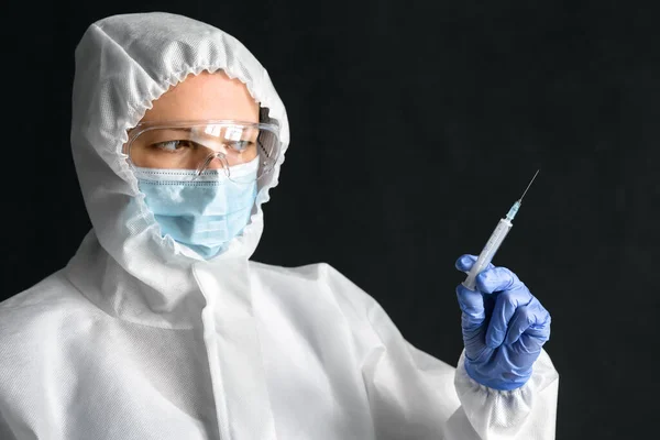 Nurse in personal protective equipment (PPE) holds syringe for COVID-19 vaccine injection. Portrait of doctor wearing face mask and goggles on dark background. Concept of PPE suit and coronavirus.