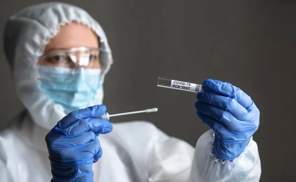 COVID-19 swab collection kit in doctor hands, nurse in personal protective equipment (PPE) suit holds tube of coronavirus PCR test. Concept of corona virus testing and diagnostics during pandemic.