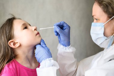 COVID-19 PCR testi ve çocuk, hemşire sevimli çocuktan alınan burun örneği için örnek tutuyor. PPE kostümlü kadın doktor, koronavirüs salgını sırasında küçük tatlı bir kızı muayene ediyor. Corona virüsü testi kavramı.