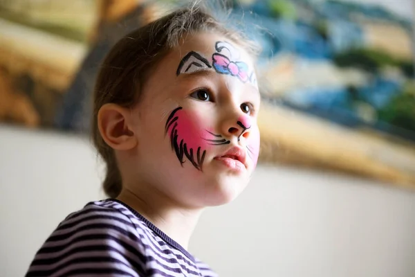 Kid with face painting of kitty, cute little girl with painted mask on face at home. Portrait of pretty child with beautiful makeup in interior, unique facial body art for party.