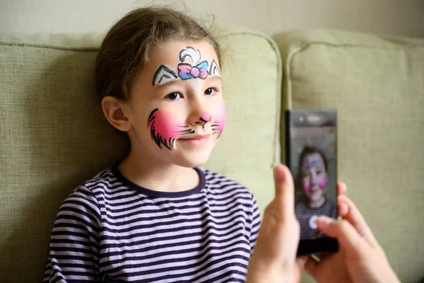 Kid with face painting of kitty, cute little girl with painted mask on face poses for photo. Happy pretty child with beautiful makeup as drawing. Adorable preschooler and unique facial art for party.