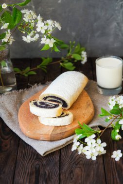 Tahta bir masada haşhaş tohumu rulosu. Ev yapımı pastalar, kiraz çiçekleri. Lezzetli tatlı ve bir bardak süt.