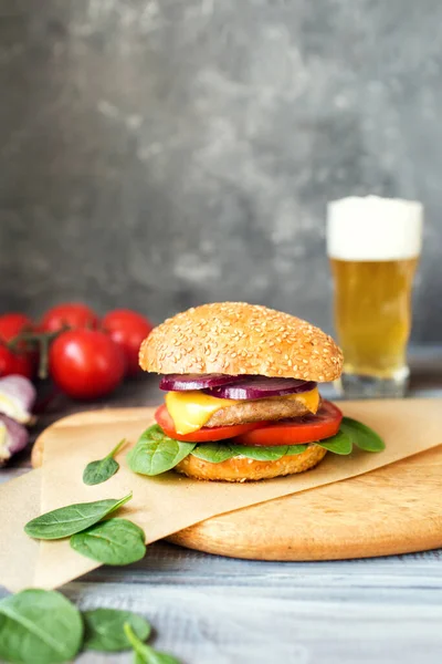 American cuisine. Burger with tomatoes, spinach and cheese. American food on a dark background. Delicious hamburger and cold beer. Space for the text. Favorite food in the USA. Cutlet on a sesame bun