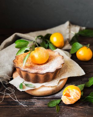 Tahta bir masada mandalina keki. Kahverengi arka planda turunçgiller. Portakallı pasta. Yapraklı mandalina. Üzerine pudra şekeri serpiştirilmiş ev yapımı kek..