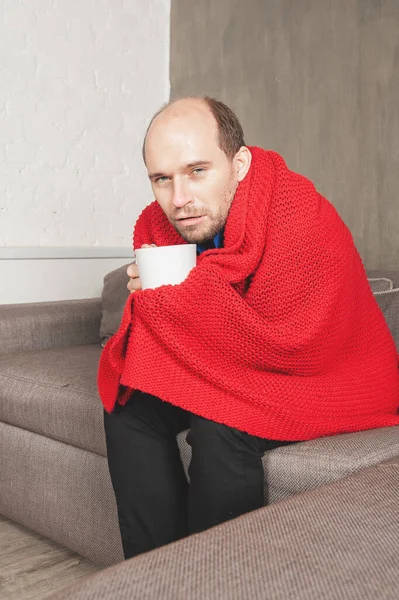 生病的男人 拿着一杯热饮 裹着温暖的针织红格子花包在客厅里 — 图库照片