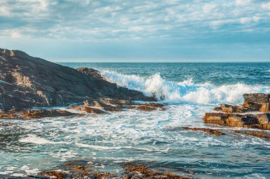 Deniz dalgaları güneşli havada kayalara çarpıyor.