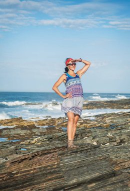 Elbiseli güzel bir kadın deniz dalgalarına karşı büyük kaya parçalarının üzerinde duruyor.