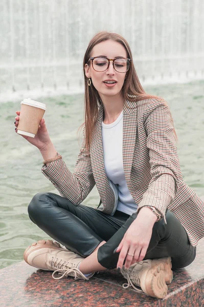 噴水の近くのパレットに座ってコーヒーを飲みながら美しいファッションスタイリッシュな女性背景 — ストック写真