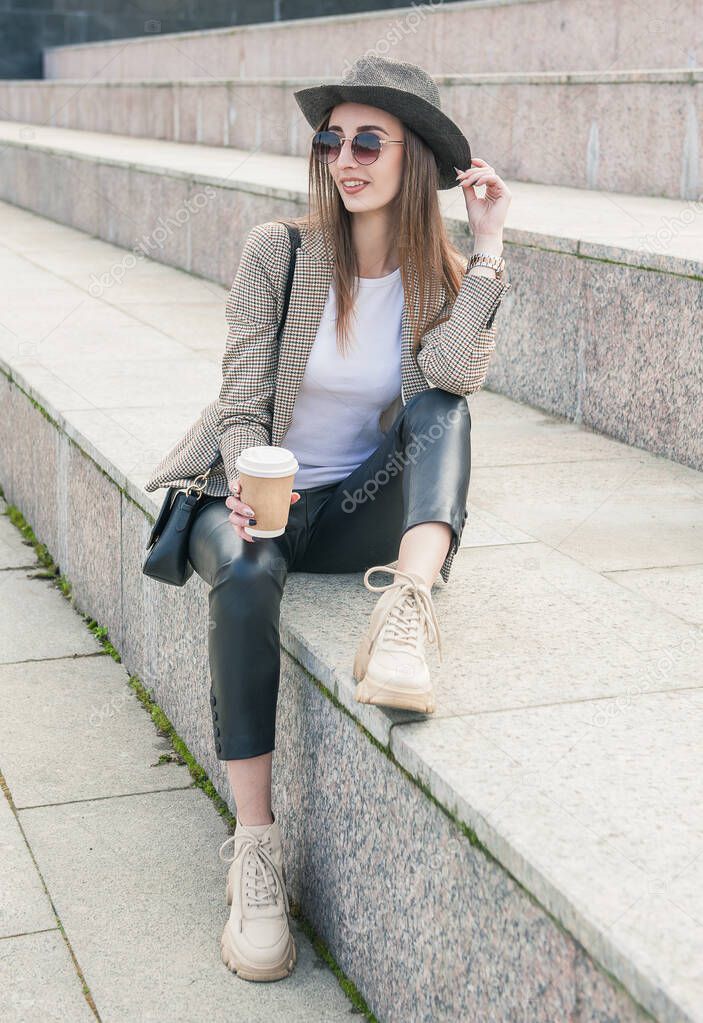 Beautiful young fashion stylish woman with cup of coffee sitting on the steps city background