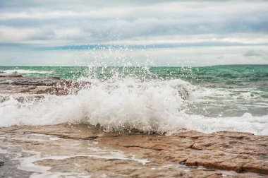 Deniz dalgaları, kayalık deniz kıyıları ve mavi bulutlarla dramatik bir sahne.