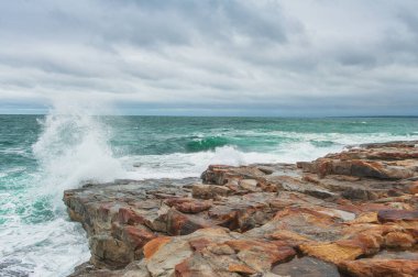 Deniz dalgaları, kayalık deniz kıyıları ve mavi bulutlarla dramatik bir sahne.