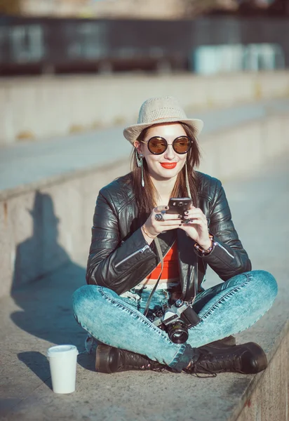 Хипстерская девушка в кожаной куртке, шляпа с ретро-камерой и мобильным — стоковое фото