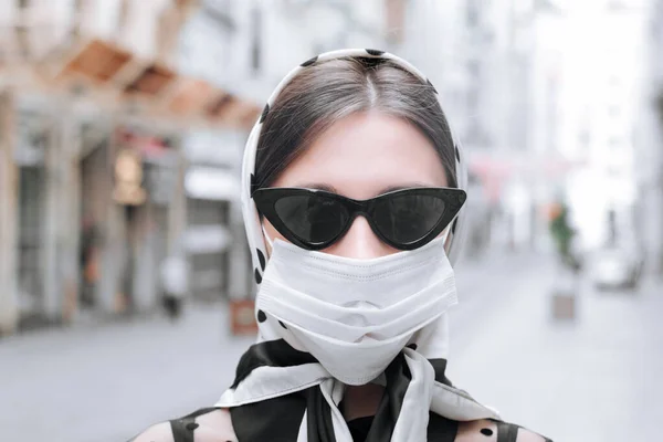 Attraktiv flicka i halsduk på huvudet och en medicinsk mask på gatan i en europeisk stad. Lady promenader på offentlig plats bär prtotective mask. — Stockfoto