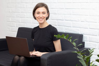 Evdeki kanepede dizüstü bilgisayarda çalışan gülümseyen genç bir kadın.