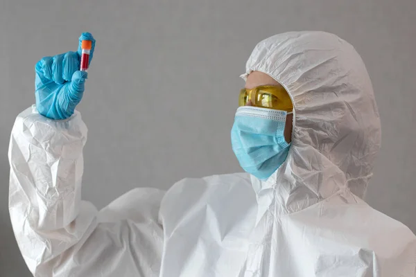 Doctor in protective suit and medical mask with gloves and yellow glasses holding a test tubes with blood and makes an analysis.