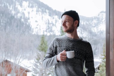 Siyah şapkalı kazaklı bir adam kış dağlarının arka planında bardak tutuyor..