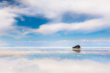 Off-road car driving on Salar de Uyuni salt flat in Bolivia. Sky with white clouds reflected in the water surface. 