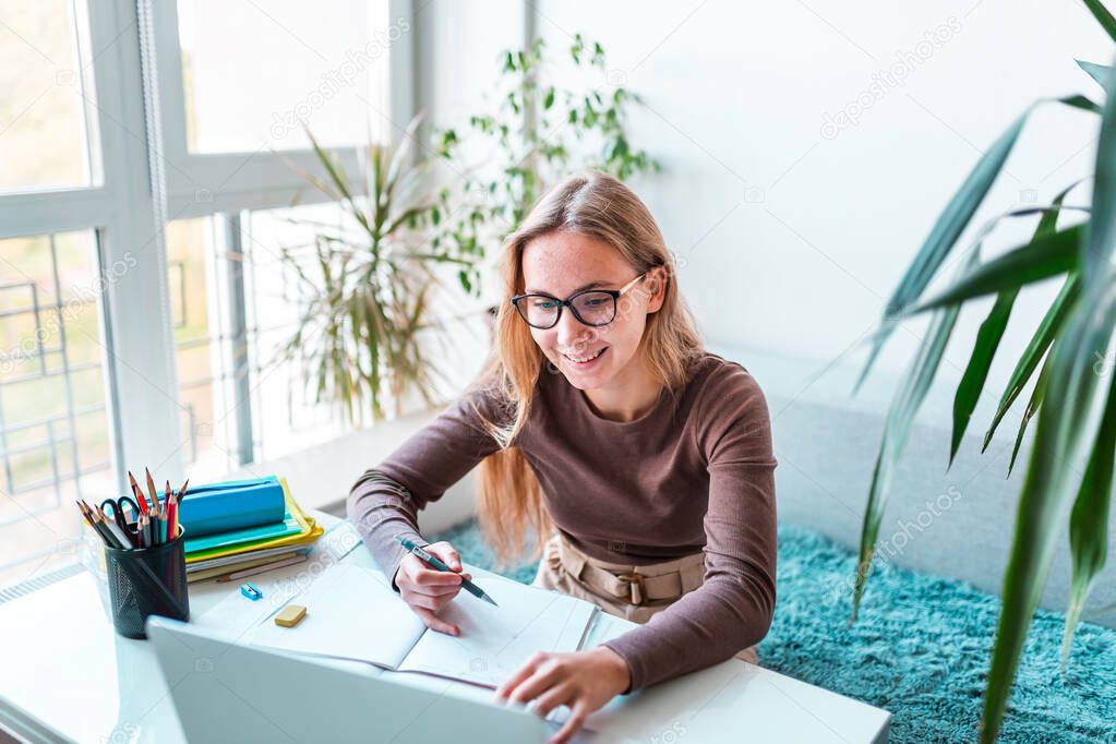 Happy beautiful young school girl left-handed working at home in her room with a laptop and class notes studying in a virtual class. Distance education and learning, e-learning, online learning concept during quarantine