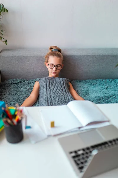 Tired and bored beautiful young school girl left-handed working at home in her room with a laptop and class notes studying in virtual class. Distance and online education, e-learning during quarantine