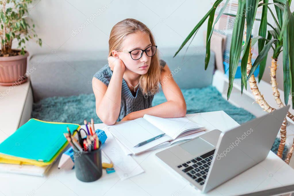 Tired and bored beautiful young school girl left-handed working at home in her room with a laptop and class notes studying in virtual class. Distance and online education, e-learning during quarantine