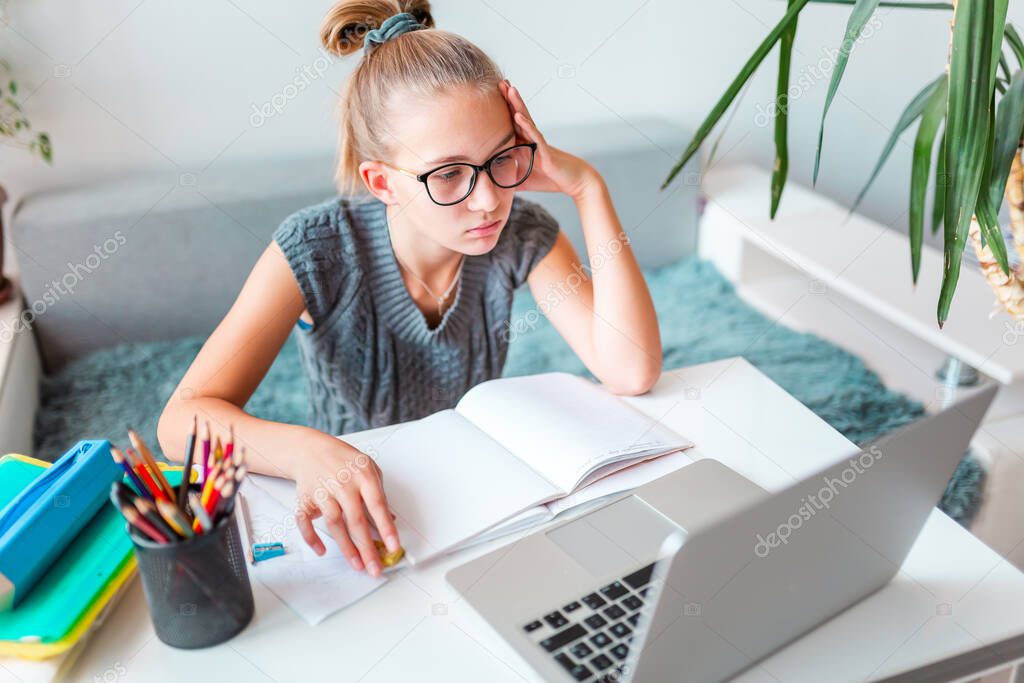 Beautiful young school girl left-handed working at home in her room with laptop and class notes studying in a virtual class. Distance and online education, e-learning during quarantine