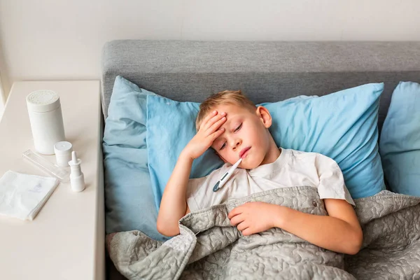 高熱と頭痛のある病気の少年はベッドに横たわって 彼の口の中で温度計を保持しています コロナウイルスの流行時に家にいて病気になったら — ストック写真