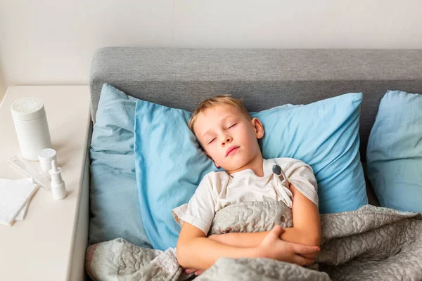 高熱や頭痛のある病気の小さな男の子がベッドに横たわって温度計を保持します コロナウイルスの流行時に家にいて病気になったら — ストック写真