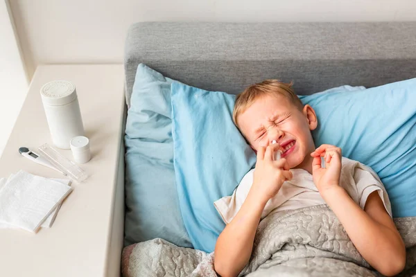 高熱と頭痛の床に横たわっている病気の小さな男の子は 彼の鼻に薬を噴霧 コロナウイルスの流行時に家にいて病気になったら — ストック写真