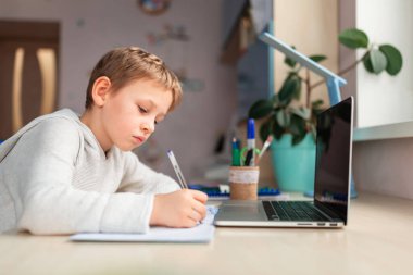Evde ödevlerini yapan sevimli bir okul çocuğu. Eğitim kitapları ve defter masanın üstünde. Uzaklık öğrenimi çevrimiçi eğitim
