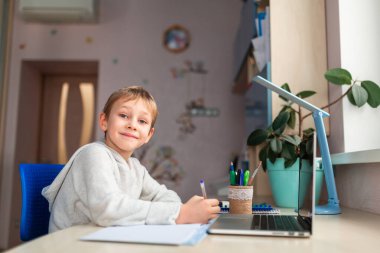 Evde ödevlerini yapan sevimli bir okul çocuğu. Eğitim kitapları ve defter masanın üstünde. Uzaklık öğrenimi çevrimiçi eğitim
