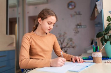 Güzel liseli kız evde ders çalışıyor. Ödev yapıyor. Eğitim kitapları ve defter masanın üstünde. Uzaklık öğrenimi çevrimiçi eğitim