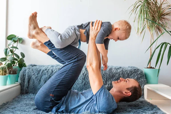 快乐的小男孩在父亲的怀里飞舞 可爱的爸爸抱着可爱的小孩儿玩飞机 在沙发上玩得开心 — 图库照片